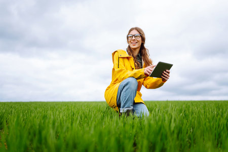 Young farmer woman in a green field, intently using a tablet. She wears a yellow jacket and glasses