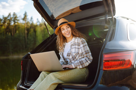 Woman sits on trunk of car, using her laptop while enjoying tranquility of a lake. Remote work.