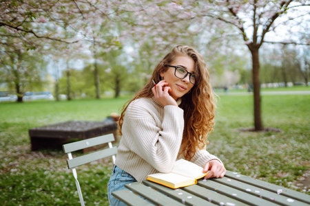 Cute woman in glasses writes in notepad, sits with laptop enjoying sunny spring day. Relax concept.