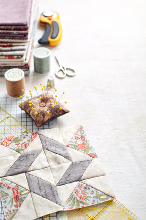 Patchwork block on craft mat, stack of fabric, sewing accessories on white wooden surface