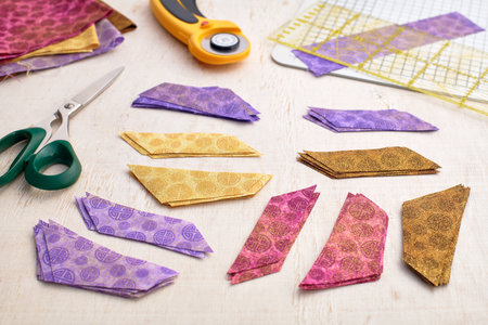 Stacks of multi-colored pieces of fabric, scissors, rotary cutter, cutting mat, ruler on a white...