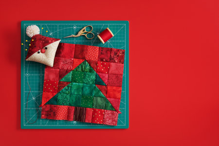 Christmas tree patchwork block, pincushion like Santa, scissors, spool of thread on craft mat, red backgroundの写真素材