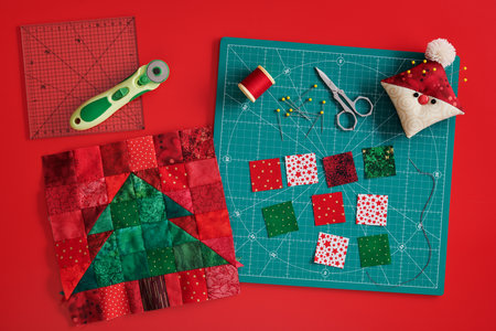 Christmas tree patchwork block, craft mat, bright square pieces of fabric, pincushion like Santa and quilting accessories on red backgroundの写真素材