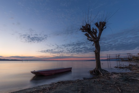 Beautiful view of Trasimeno lake (Umbria) at dusk, with a little boat near a bare tree, perfectly still water and a mackerel skyの写真素材