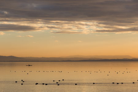 Beautiful view of a lake at sunset, with orange tones, birds on water and a man on a canoeの写真素材