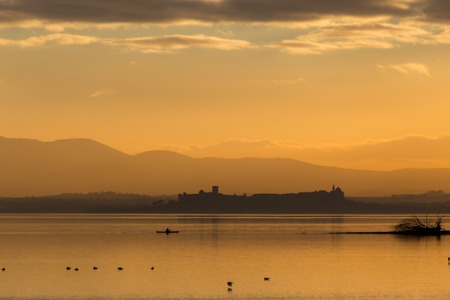 Beautiful view of Trasimeno lake (Umbria, Italy) at sunset, with orange tones, birds on water, a man on a canoe and Castiglione del Lago town on the backgroundの写真素材