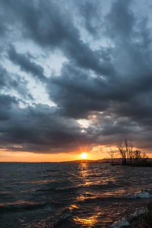 Beautufl sunset on a lake, with tree silhouettes and sun coming down under an overcast skyの写真素材