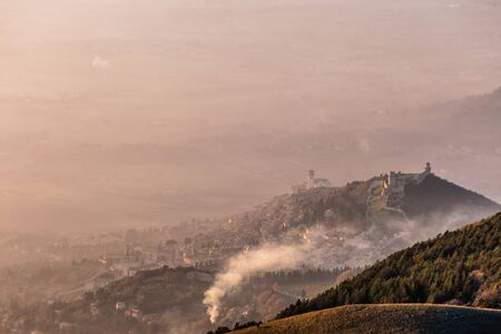 A view from above of Assisi town (Umbria, Italy) in the middle of mist, with beautiful, warm sunset colors.の写真素材