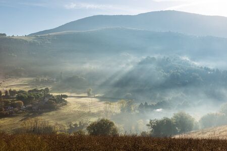 Sunrays at dawn above hills cutting through the mist and trees.