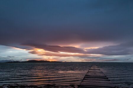 View of a pier on a lake with a ray of sun against a moody skyの写真素材