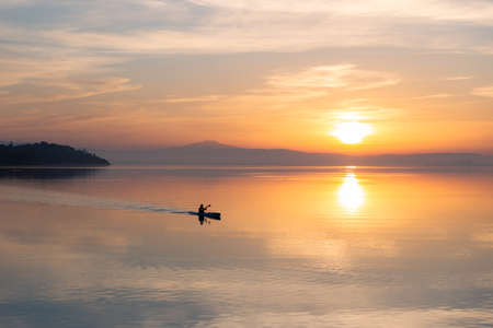 Beautiful sunset at Trasimeno lake with a man on a canoe above perfectly still waterの写真素材