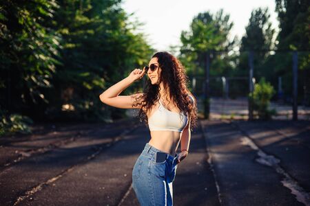 A young brunette in glasses with long curly hair is standing in the street in a white top and jeans,...