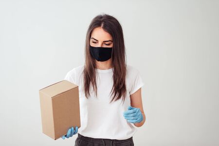Cute girl in a mask and gloves looks at a cardboard box, package delivery. Safe delivery during the outbreakの写真素材