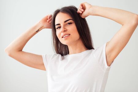 Gorgeous European woman smiling, keeping her hands behind your head. nice brunette in a clean white t-shirt on a light background.の写真素材