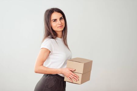 The brunette stands sideways in a white shirt with a box in her hands and smiles sweetly. Sending small boxes by mail.の写真素材