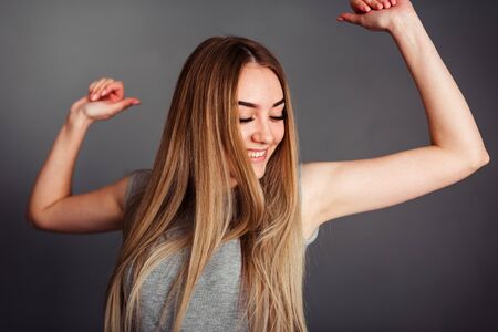 portrait of a young Caucasian blond haired woman in a gray T-shirt, cheerfully looking to the side, raising her hands while dancing, listening to music that is in a good mood and happy on a gray wallの写真素材