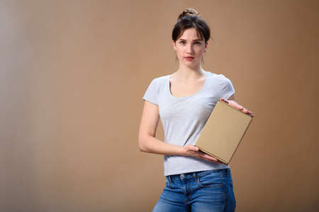 Cute girl in a gray T-shirt holds a craft box in her hand with an edge to the camera. And shows the class with a thumb up on a beige backgroundの写真素材