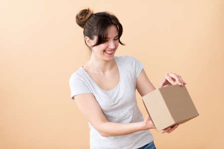 Cute girl in a gray T-shirt holds a craft box in her hand with an edge to the camera. And shows the class with a thumb up on a beige backgroundの写真素材