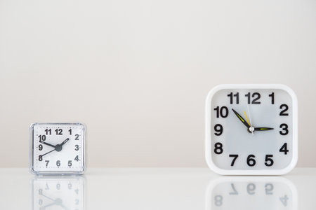 There are two clocks on the table. Different times, big clock on the right, small clock on the left. On the table a mirror reflection of a white backgroundの写真素材