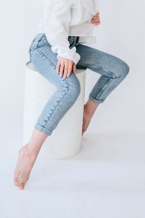 A girl in jeans and a white shirt sits casually on a white barrel. Close up photo of female legs in jeansの写真素材