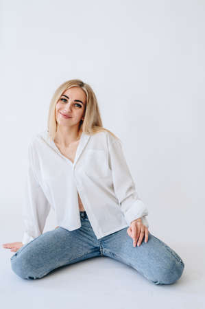 A girl in jeans and a white shirt sits casually on her knees. Blonde woman on a white background in...