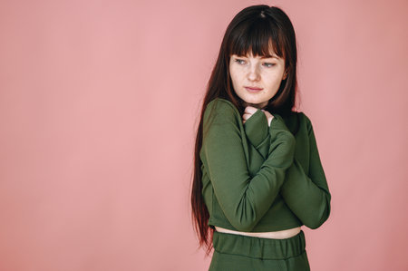 A young girl on a pink background stands in a closed pose with her arms crossed over her chest and...