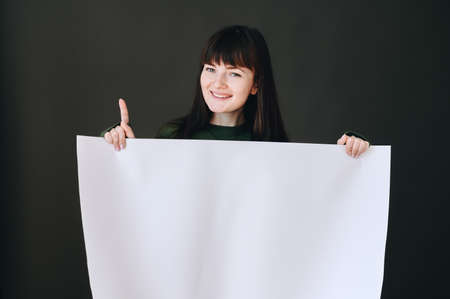 Horizontal photo of a girl who holds a white paper in her hands and points up with her index finger....