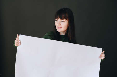 A young girl on a black background holds a large sheet of clean white paper and looks to the side. Concept of free space for advertisingの写真素材