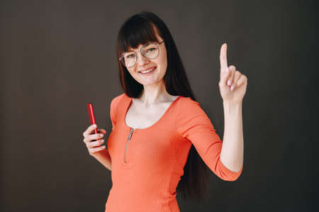 A teenager with a phone in his hands and glasses smiles broadly and shows his index finger up.
