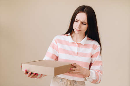 A frowning girl attentively holds a flat box in her hands on a beige background in the studio. Girl...