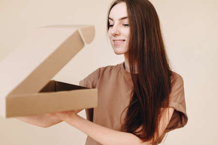 Smiling brown-haired woman looks into the box and sniffs food, wearing a brown t-shirt on a beige background. The girl received her long-awaited delivery.の写真素材