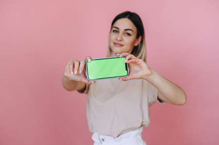 girl is holding horizontally a smart phone with a green screen on a pink background, focus on the phone. Place for advertisingの写真素材