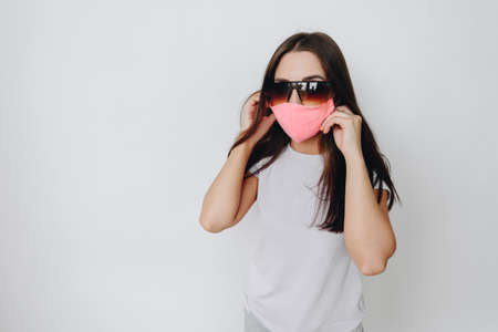 A woman in a white t-shirt during quarantine due to a flu outbreak. The model is dressed in a protective stylish handmade mask, large sunglasses on a white isolated backgroundの写真素材