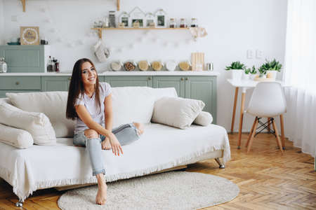 A girl in a T-shirt and torn jeans sits on the couch with one leg bent and looks at the camera. The...