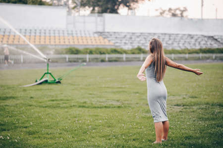 A girl in a gray dress stands with her hands apart and waits. A lawn sprinkler sprays water in a stadium. Watering the grass on a summer eveningの写真素材