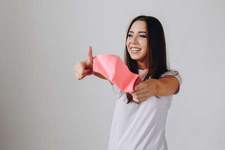 A girl in a white T-shirt on a white background holds a face mask in front with two hands and looks away with a smile. The concept of health protection.の写真素材
