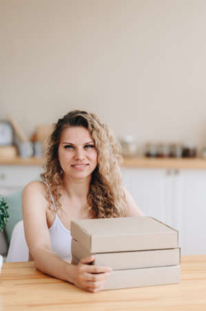 A vertical photo of a curly-haired girl sitting at a table at home, a stack of boxes on the table, the girl arranged the parcels. Sunny day. copy spaceの写真素材