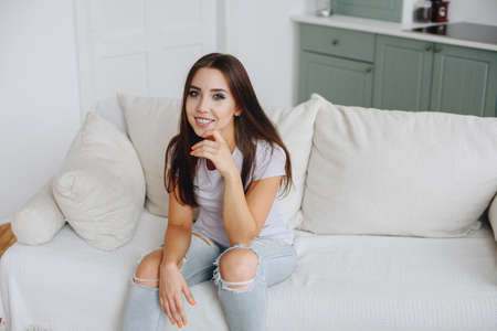 Happy brunette girl in a white t-shirt sits on the sofa, relaxes. Concept Rest at home