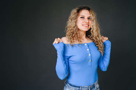 The girl raises her arms in her elbows up against the background of a dark wall. Curly-haired girl in a blue sweater. Concept natural girl without makeup.の写真素材