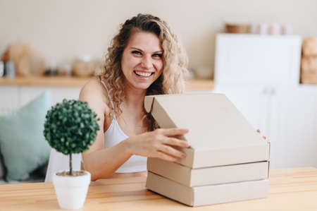 The girl sits at the table at home and opens a flat box. Curly blonde in a good mood. Empty space for text. The concept of receiving a surprise mail.の写真素材