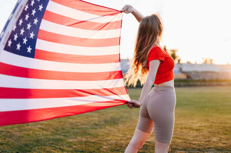An American Flag athlete with an American Flag on Sunset develops on Wind. Woman in Red Top Holds Flag and Looks at Sun. girl at stadiumの写真素材