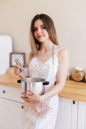 Young Woman sexy in an Apron Holds Saucepan in Her Hands and Stirs with Whisk. Vertical Portrait of Housewife. Good Morning Prepare Breakfast.の写真素材