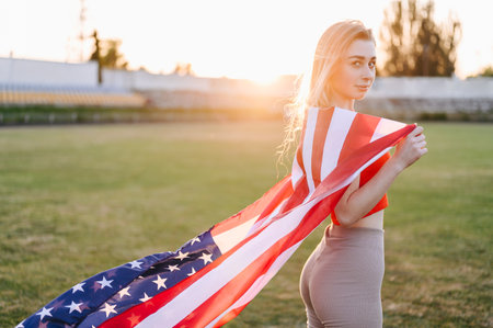 Athletic Thin Woman Stands Against The Sun And Holds The American Flag On Her Shoulders. close-up portrait. Summer Evening in Nature.の写真素材