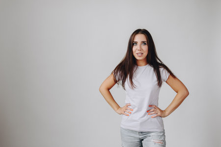 Girl with Strange Grimace Stands on White Background. Woman in White T-Shirt and Hands on her hips. Remember Error. Forgot, Bad Memory Concept.の写真素材