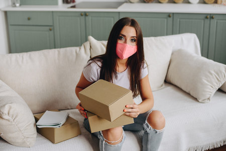 Photo From Above, Girl in Pink Mask Sits On Sofa with Boxes in Hands. Girl during Quarantine Works at Home. Soft Light sofa with Pillows.の写真素材