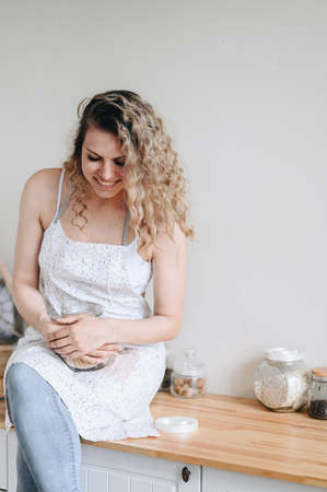 Vertical photo Woman in Apron Sits on Wooden Table and Laughs. Cozy Home Kitchen. Prepare a delicious meal.の写真素材