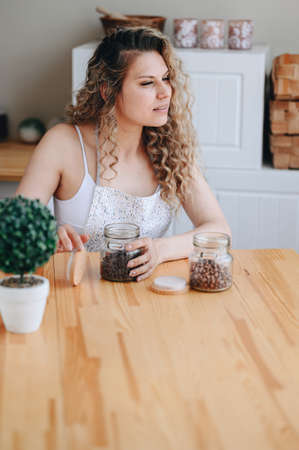 Curly Haired Woman sits at Wooden Table and Looks Away. Coffee Beans in Glass Jar on Table. Girl is Sitting in Kitchen in Morning.の写真素材