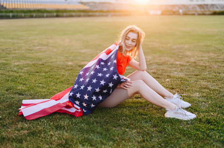 Girls with American Flag on their Shoulders are sitting on Grass. Girl Sheltered with Flag and Closed Her Eyes from Pleasure. Memorial Day Sports Girl. Sunset at Golden Hour.の写真素材