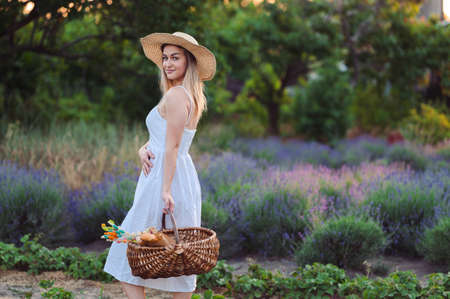 Young Woman in White Dress and Hat Stands Among Lavender and Strawberries. Summer Evening in Village. Enjoy Life on Vacation.の写真素材