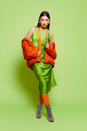 High fashion portrait of young Asian woman on light green background. Orange down jacket, orange tights, light green dress, orange tieの写真素材